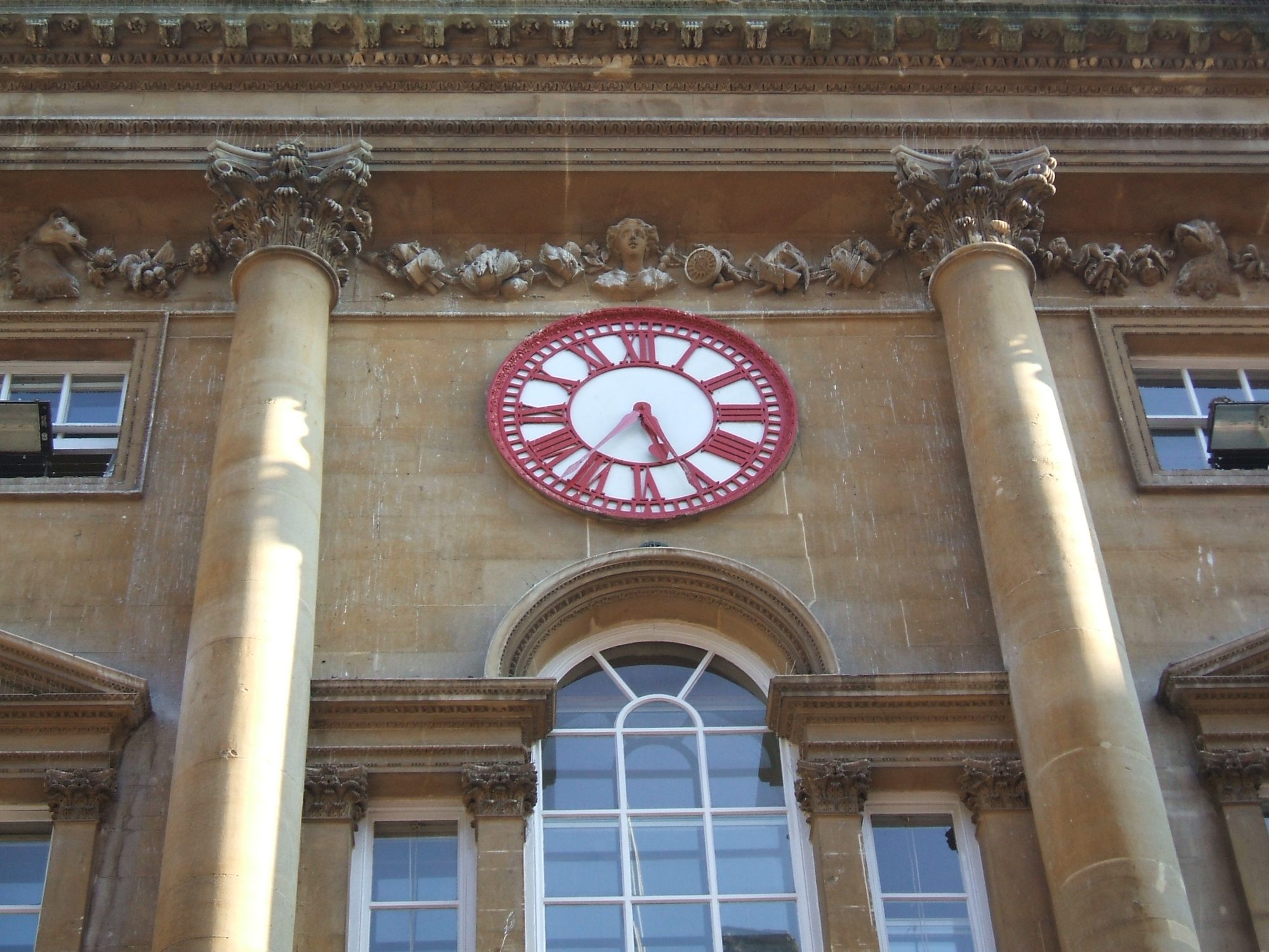 A large red-and-white clock face mounted between stone columns on the Georgian front of the Exchange in Bristol, with two minute hands