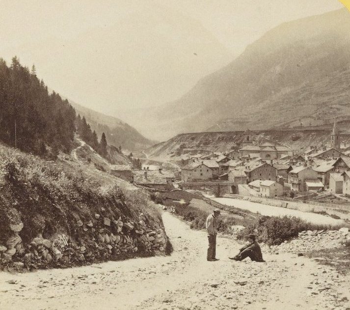 An old photograph of the village of Lanslebourg at the foot of the Mont Cenis road, with two men resting by a dusty track and mountains behind