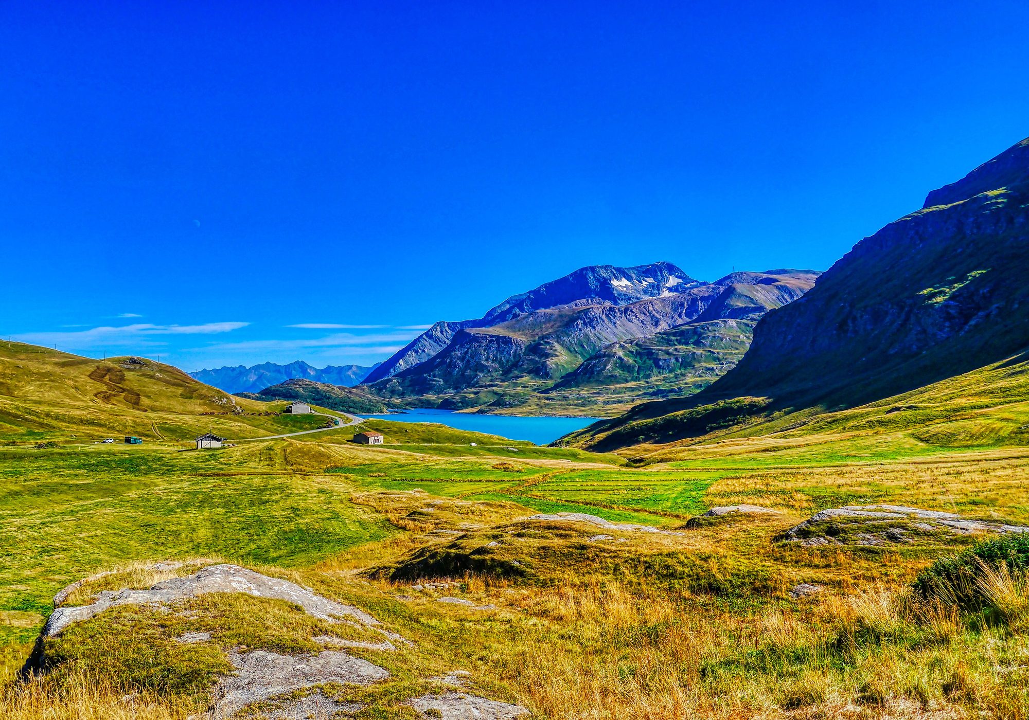 The green high plateau of the Col du Mont-Cenis under a blue sky, with the lake and snow-patched peaks in the distance