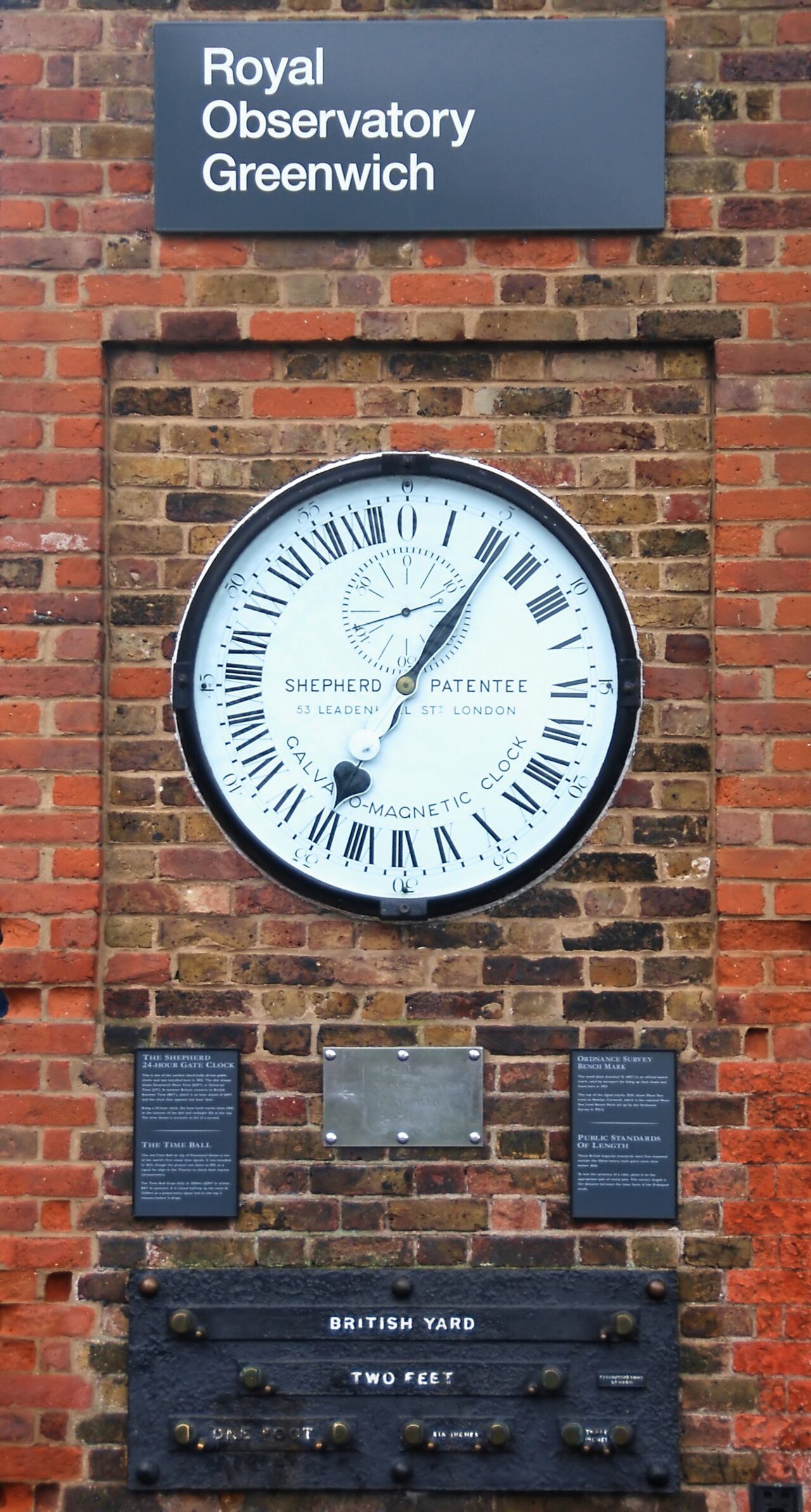 A large round twenty-four-hour clock set into a red brick wall beneath a sign reading Royal Observatory Greenwich, with British standard measures of length mounted below it